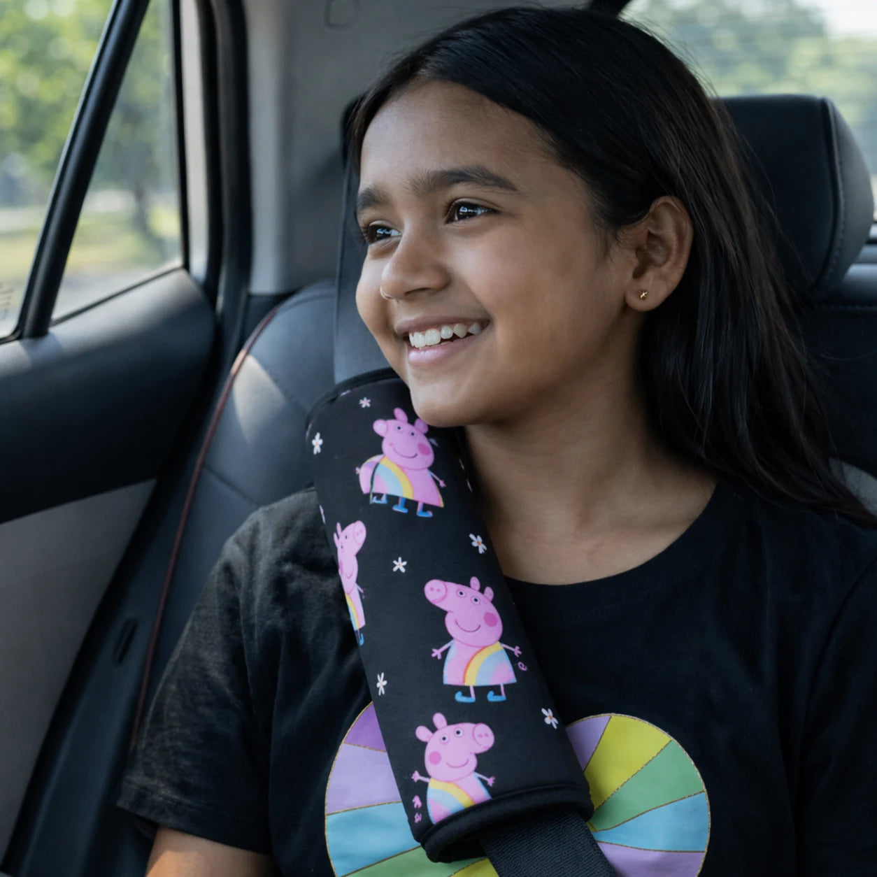 A young Indian girl smiling in a car while wearing a Car-Wah! black kids' seatbelt cover featuring a colorful Peppa Pig pattern. Making traffic a little more fun with Car-Wah! comfort car accessories.