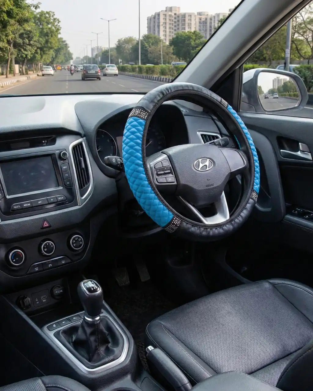 Blue PU leather steering wheel cover with anti-slip rhinestone grip installed in car interior during city drive. A premium Car-Wah! car accessory to show off your ride's personality.